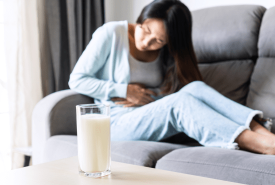Femme assise sur un canapé en arrière-plan, semblant éprouver un léger inconfort abdominal, tandis qu’un verre de lait est posé au premier plan sur une table. L’image illustre les symptômes digestifs liés à une intolérance au lactose