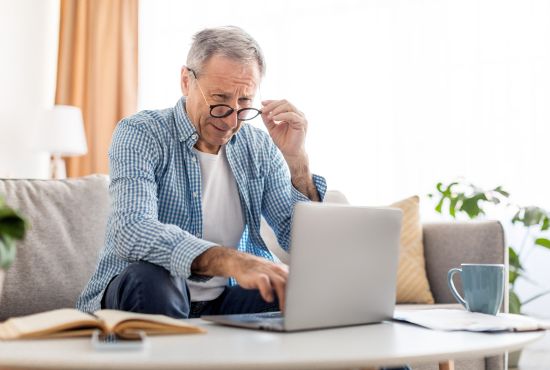 Une personne âgée est assise dans son fauteuil, devant son ordinateur. Elle porte des lunettes qu'elle abaisse légèrement, visiblement gênée par sa vision.