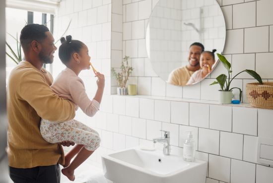 Un père tenant sa fille dans ses bras dans une salle de bain, l'aidant à se brosser les dents devant un miroir, illustrant les bons réflexes pour la santé bucco-dentaire des enfants.