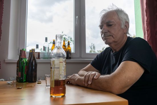 Un homme âgé assis à une table entourée de nombreuses bouteilles d'alcool vides, regardant un verre devant lui avec une expression pensive, illustrant les signaux précoces d'une consommation problématique d'alcool.