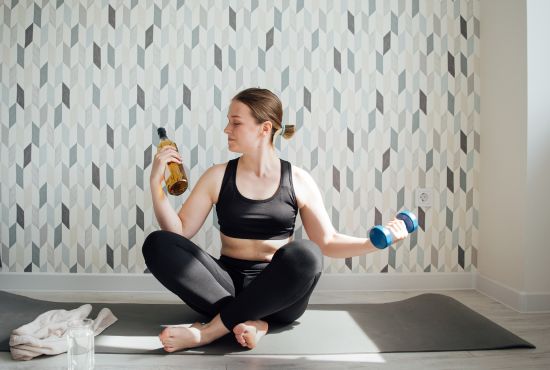 Une femme en tenue de sport assise sur un tapis de yoga, tenant une bouteille de bière dans une main et un haltère dans l'autre, illustrant le conflit entre la consommation d'alcool et les performances sportives