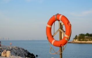 Une bouée de sauvetage circulaire rouge et blanche installée sur un poteau en bord de mer, sur fond de ciel clair et d'eau calme.