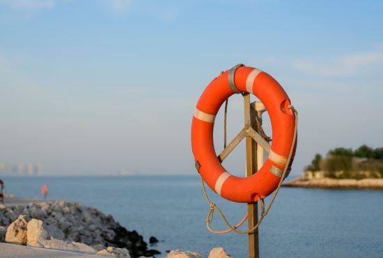 Une bouée de sauvetage circulaire rouge et blanche installée sur un poteau en bord de mer, sur fond de ciel clair et d'eau calme.