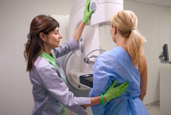 Une manipulatrice en radiologie positionne une patiente vêtue d'une blouse bleue devant un mammographe pour réaliser un examen du sein.