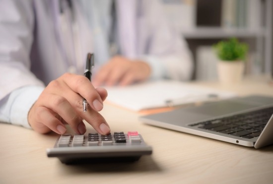 Un professionnel de santé en blouse blanche utilisant une calculatrice à son bureau pour estimer le coût et le remboursement de soins médicalisés.