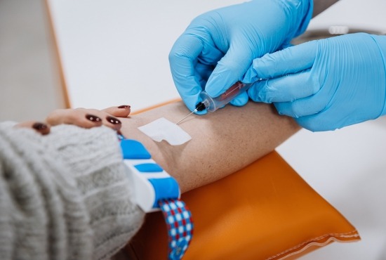 Un professionnel de santé portant des gants bleus effectue un prélèvement sanguin au niveau du bras d'un patient équipé d'un garrot.