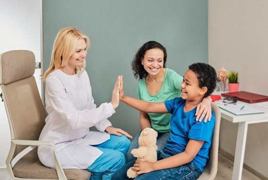 Une médecin pédopsychiatre en blouse blanche tape dans la main d'un jeune garçon souriant assis aux côtés de sa mère dans un cabinet médical.