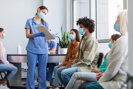 Une professionnelle de santé en blouse bleue et masque médical accueillant des patients masqués dans une salle d'attente pour la vaccination.